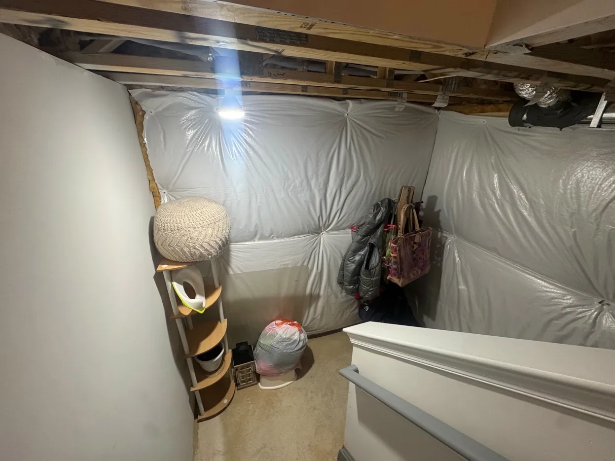A basement corner with freshly painted white walls, exposed wooden ceiling beams, plastic insulation sheeting on some walls, a small wooden shelf holding household items, hanging coats, and a white staircase railing.